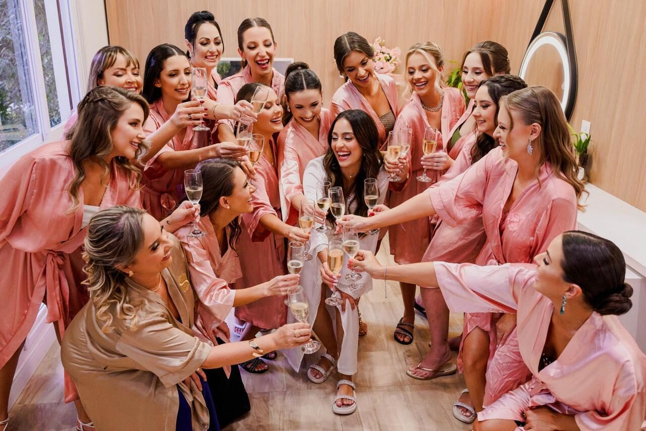 Grupo de madrinhas em robes rosé brindando com champanhe em torno da noiva na suíte — Salão do Bosque