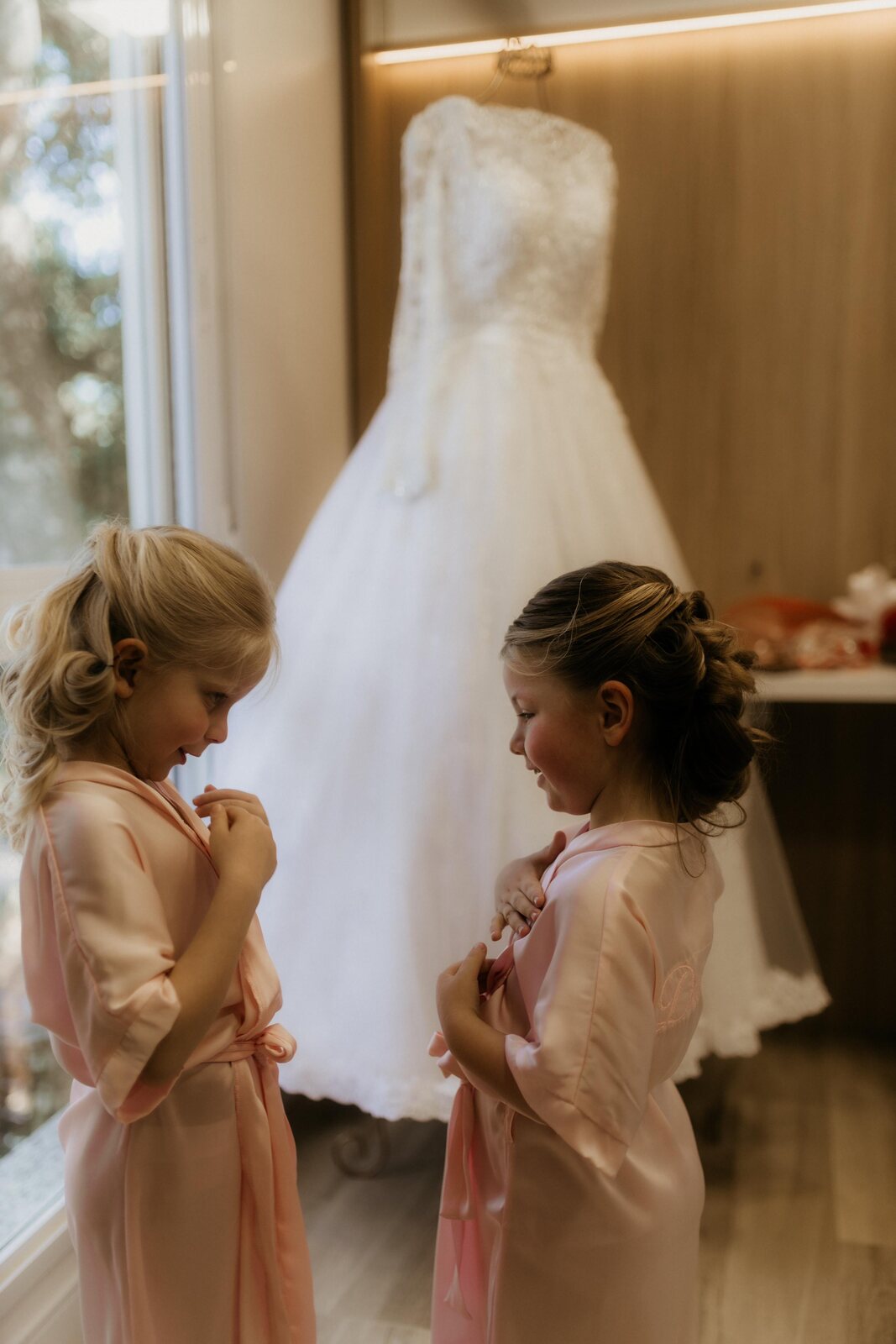 Duas daminhas em robes rosa observando o vestido de noiva pendurado — Salão do Bosque