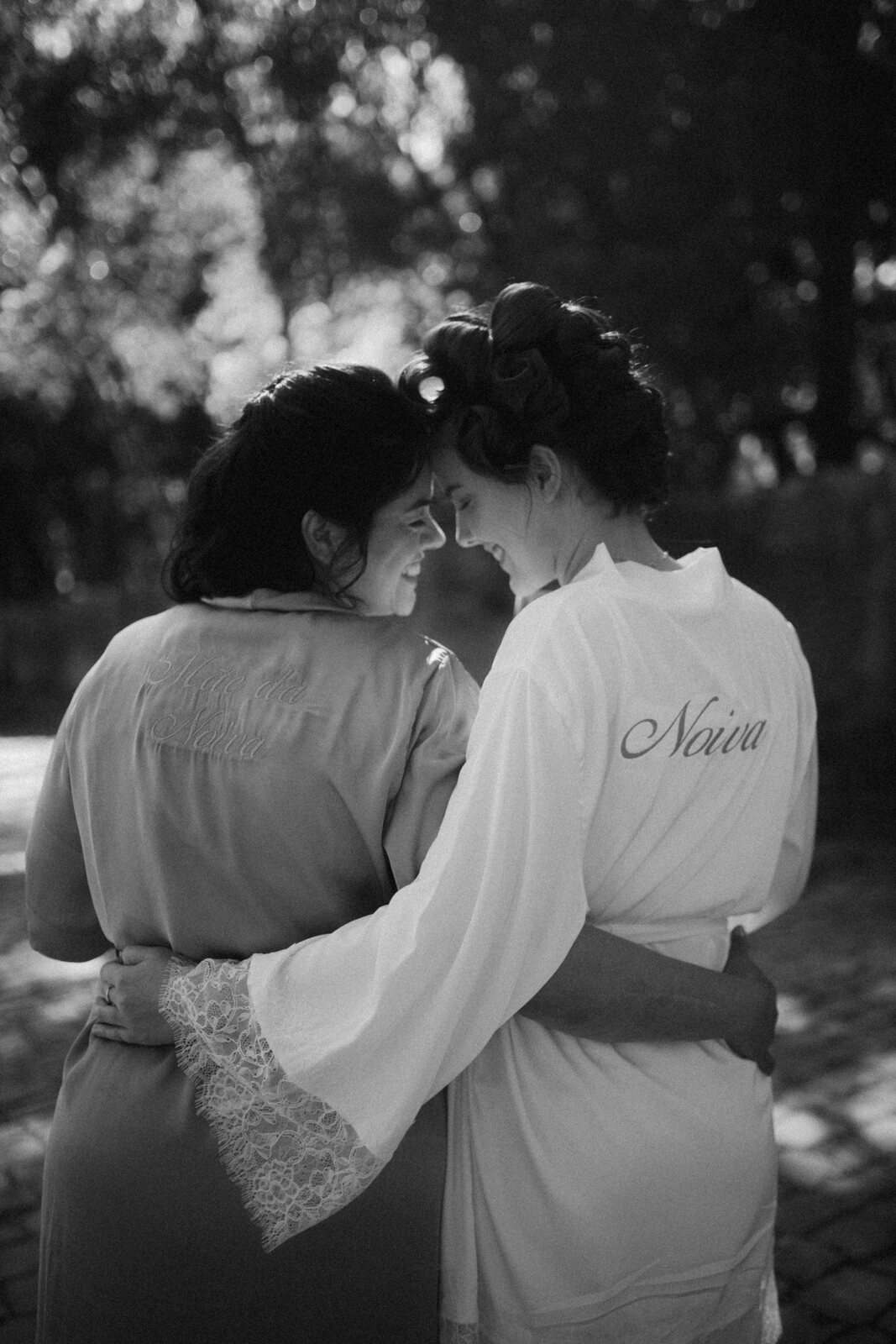 Noiva e mãe da noiva abraçadas, com robes bordados em ouro ao ar livre — Salão do Bosque